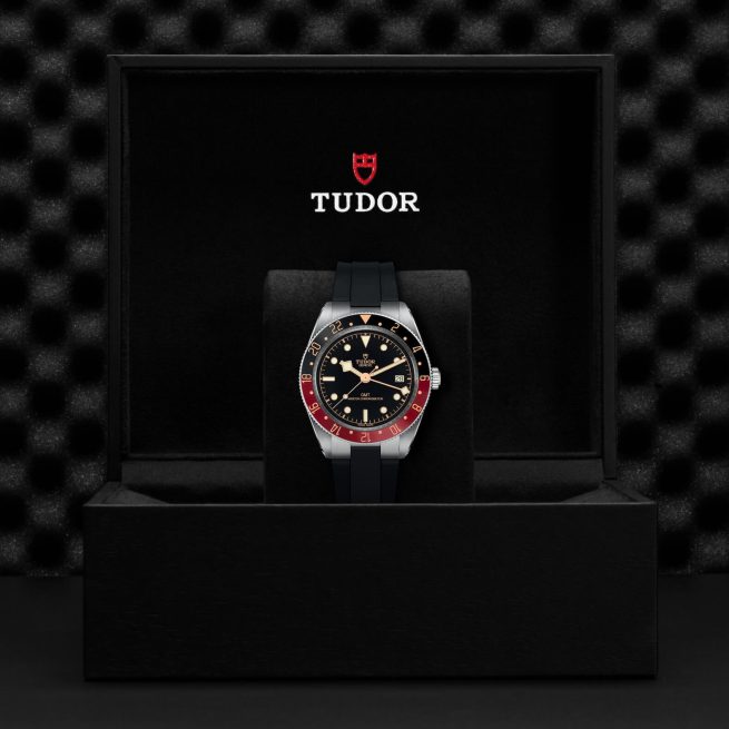 A Tudor M7939G1A0NRU-0002 watch with a black, red, and gold bezel is displayed in an open black box with "Tudor" and a red shield logo on the inner lid.