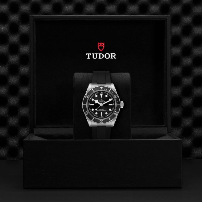 A black Tudor watch with a silver bezel and black rubber strap is displayed in an open black box with the Tudor logo inside the lid.