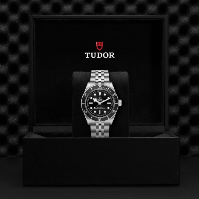 A Tudor wristwatch with a black dial and silver bracelet is displayed inside an open black box with the Tudor logo on the interior lid.