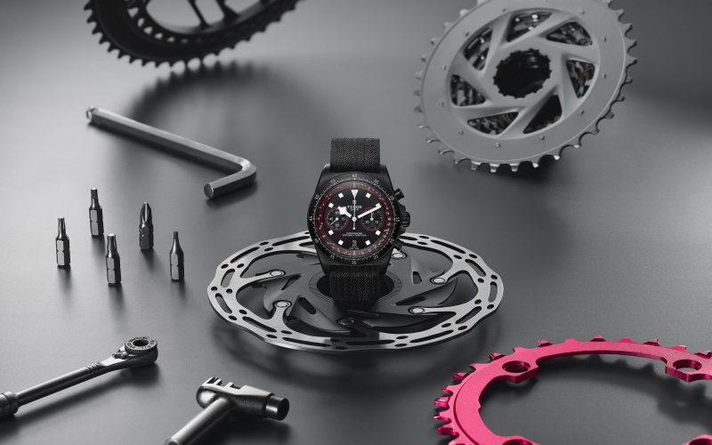 A black wristwatch with a red-accented dial is displayed among various bicycle gears, including chainrings, sprockets, and tools, on a gray surface.