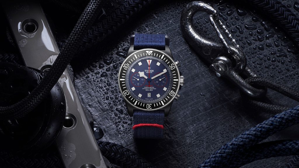A silver watch with a navy blue strap displayed on a surface with wet, black ropes and metal shackle nearby.