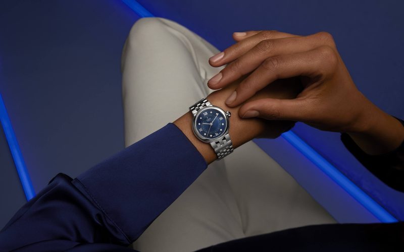 Close-up of a person's hand adjusting a silver wristwatch with a blue dial on their left wrist, against a dark background with blue accents. The person is wearing a dark blue shirt and white pants.