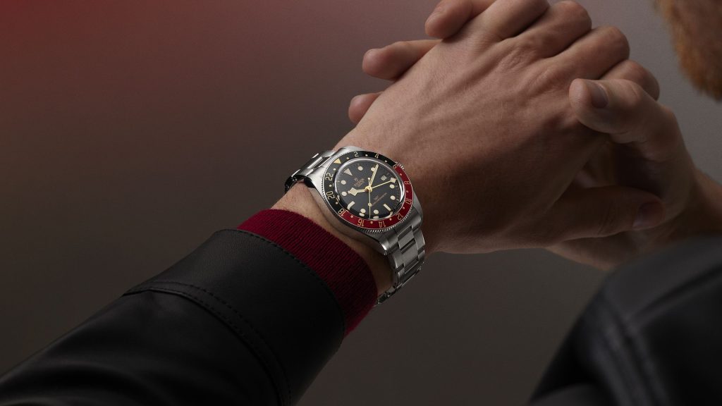 Close-up of a person wearing a stainless steel wristwatch with a black and red bezel, and a black dial with white markings, clasping their hands together.