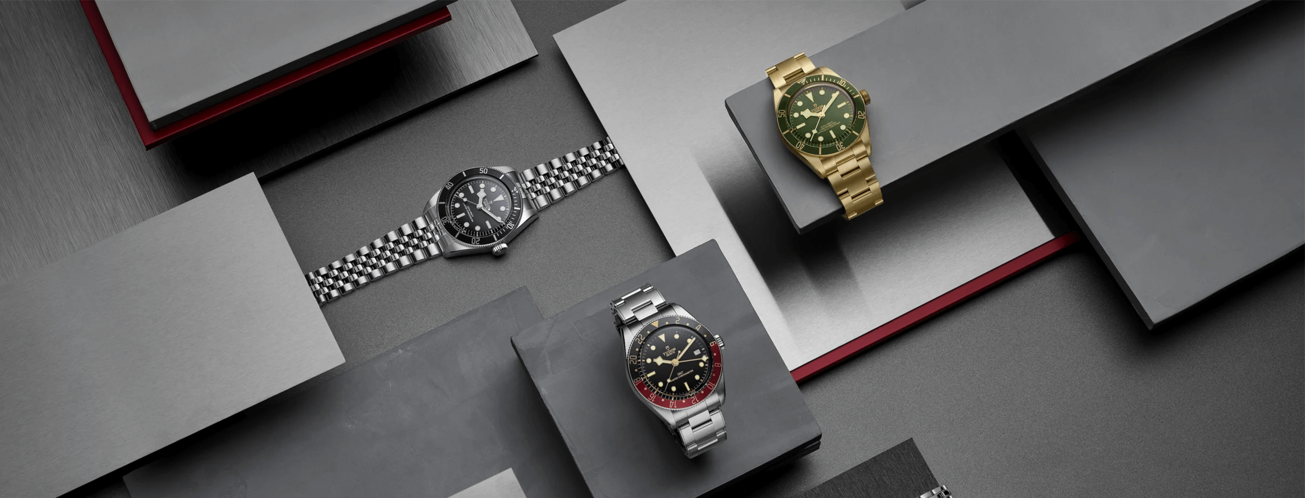 A display featuring three luxury wristwatches, with one silver-black, one gold-green, and one black-red, arranged on geometric grey platforms.