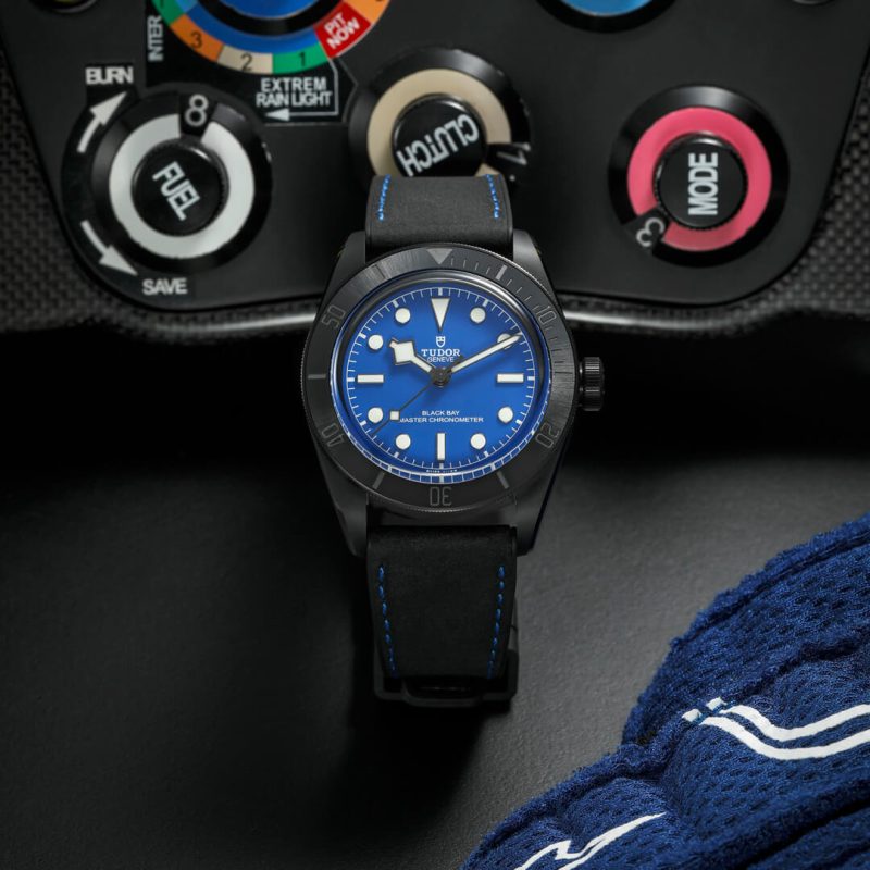 A black watch with a blue dial and blue accents is displayed next to an instrument panel and blue clothing.