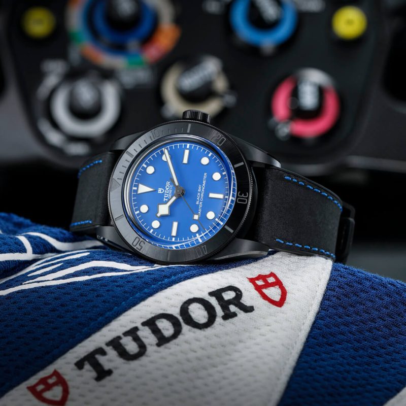 A black wristwatch with a blue dial and bezel, displayed against a blue and white cloth with "Tudor" written on it. The background shows blurred gauges and dials.