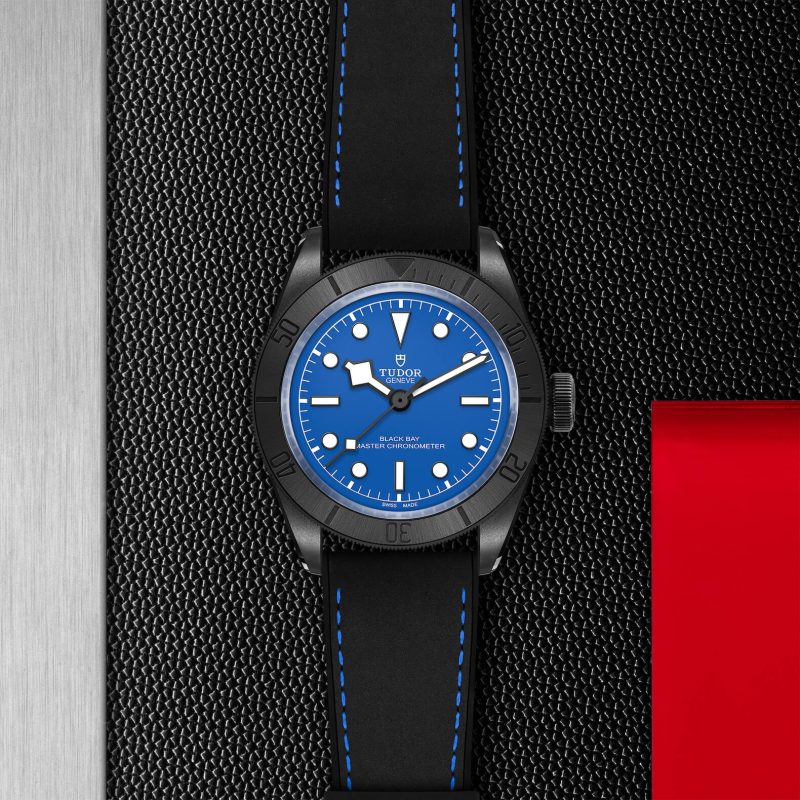 A black wristwatch with a blue dial and white markers, branded "Tudor," with a black and blue stitched strap, displayed on a textured black surface next to a red rectangular object.