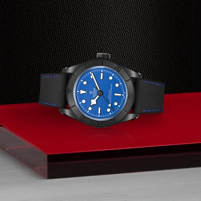 A wristwatch with a blue dial and black strap is displayed on a red and silver surface against a dark, textured background.