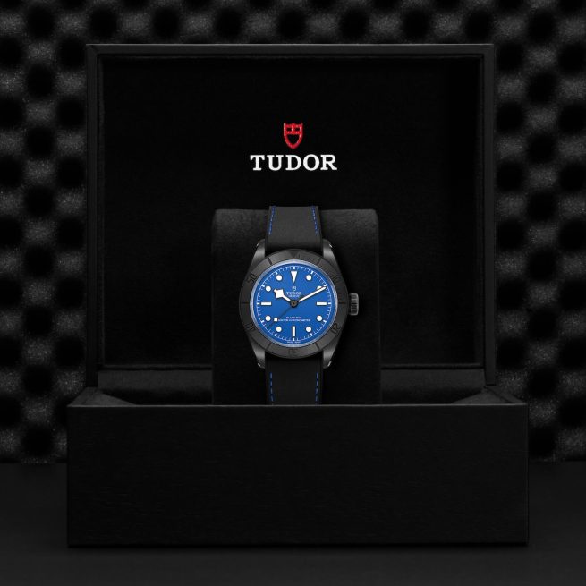 A Tudor wristwatch with a blue face and black band displayed inside an open black case with Tudor branding.