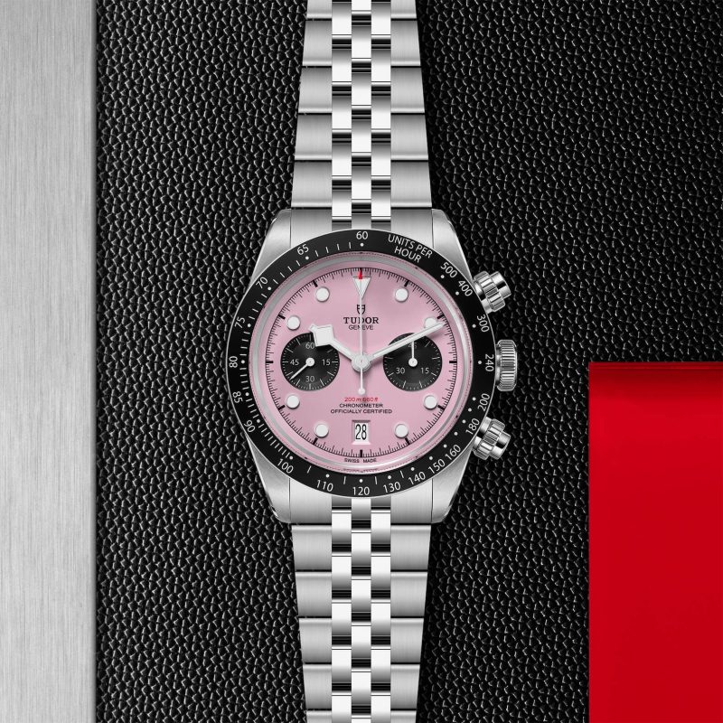 A stainless steel wristwatch with a pink dial, black sub-dials, a tachymeter bezel, and a silver bracelet, displayed against a textured black background with a red accent.