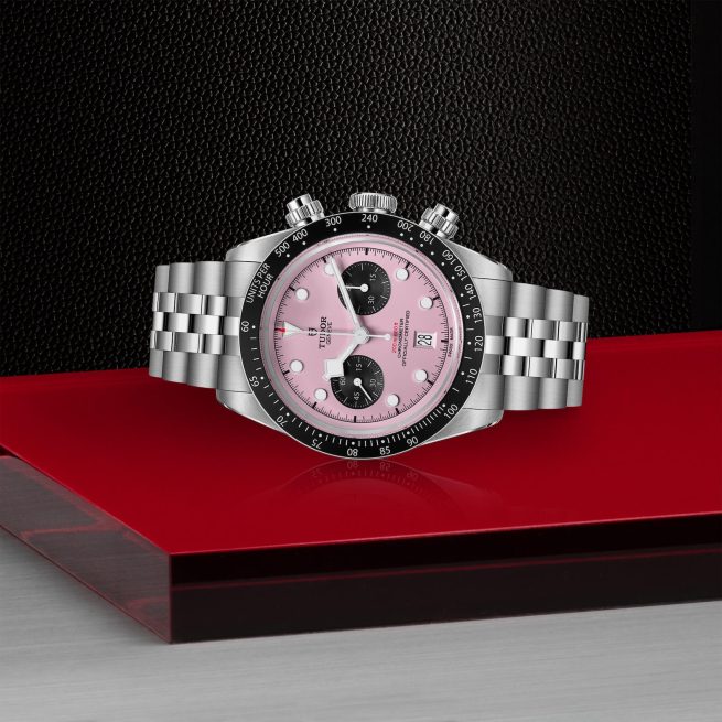 A stainless steel wristwatch with a pink dial, black subdials, and a black bezel is displayed on a red and black surface. The watch has a metal bracelet and the brand name "Tudor" is visible on the dial.