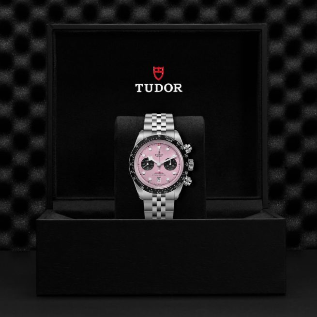 A Tudor wristwatch with a pink dial and silver band is displayed in an open black box featuring the Tudor logo.