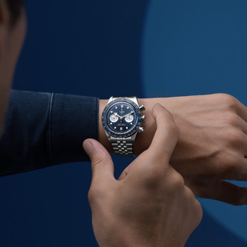 A person wearing a silver wristwatch with a blue dial and three sub-dials; their other hand is adjusting the watch's bezel against a dark blue background.