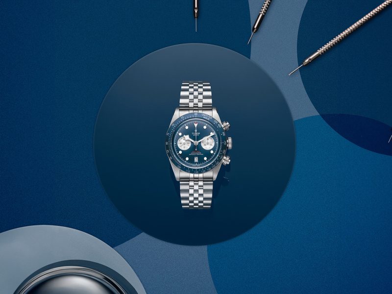 Silver wristwatch with a blue dial and silver subdials, displayed on a dark blue circular platform. Surrounded by watchmaking tools on a blue background, showcasing the artistry behind new watches.