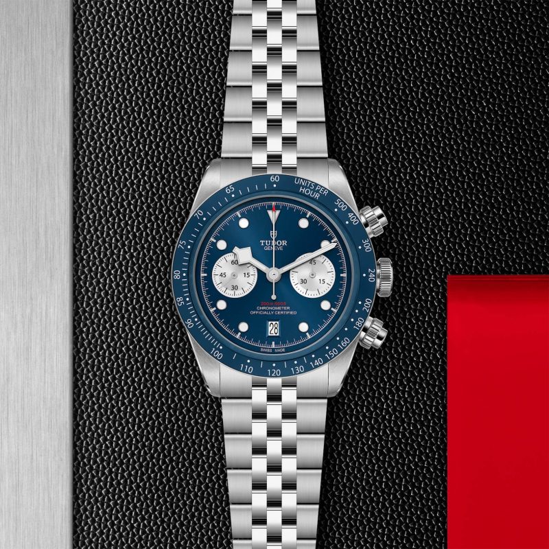 A silver wristwatch with a blue dial, white subdials, and a checkered metal band is displayed on a textured black and red surface.