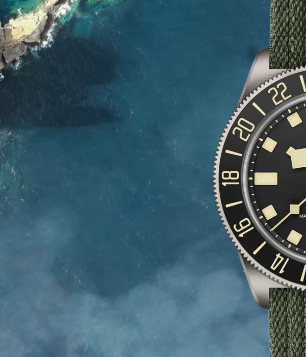 A TUDOR Pelagos FXD GMT watch with a green strap and black dial elegantly complements the aerial view of a coastline, where water meets lush greenery.