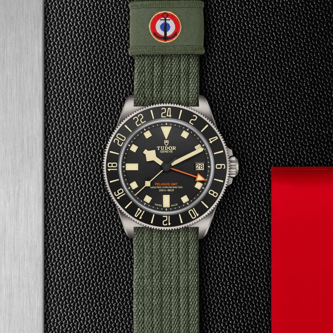 The Pelagos FXD GMT Tudor watch features a green fabric strap, a black dial with an orange hand, and a date display, all set against a textured black and silver background with a red accent.