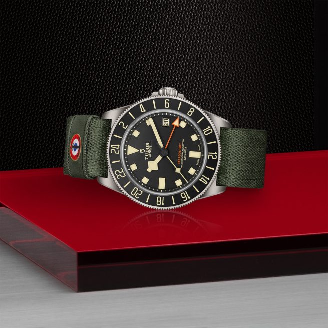 The TUDOR Pelagos FXD GMT watch, featuring a black and orange face, a green strap, and a stainless steel casing, is displayed on a red and black platform.