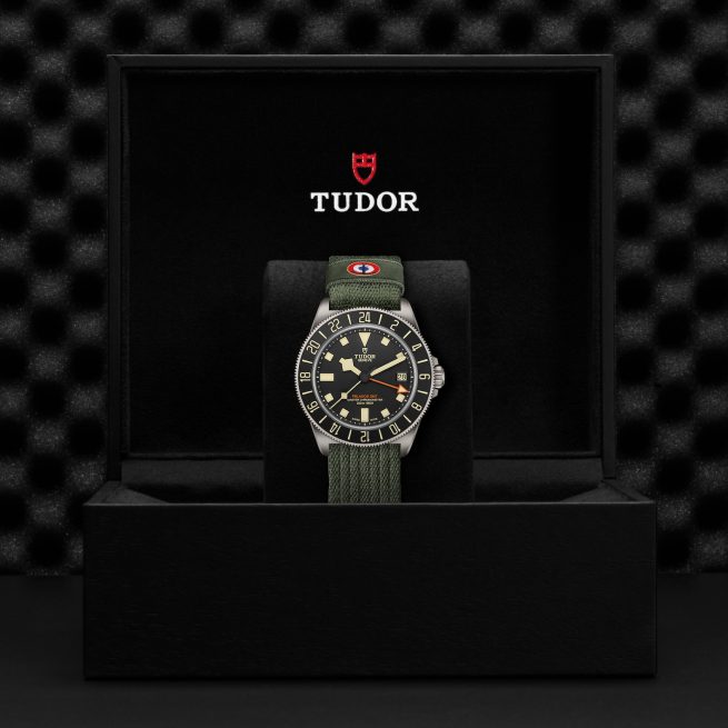 The TUDOR Pelagos FXD GMT watch featuring a green fabric strap is showcased in an open black box against a textured black background.