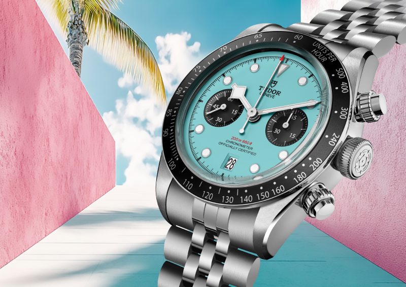 The M79360N-0024 silver wristwatch showcases a turquoise face with black dials, set against a tropical scene featuring pink walls, a palm tree, and blue sky.