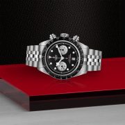 The M79360N-0013, a stainless steel wristwatch featuring a black dial with white subdials and a date display, rests on a red and black textured surface.