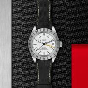 The M79470-0006 Tudor wristwatch, featuring a white dial, black bezel, and leather strap, is showcased against a textured black background with metallic and red highlights.