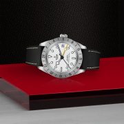 The M79470-0006 watch, featuring a silver case, white face, black strap, and yellow-tipped hand, is displayed on a red surface with a textured black background.