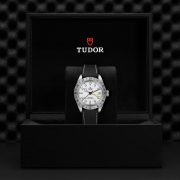 M79470-0006 comes in a black Tudor box with a red shield logo and textured interior, featuring a silver wristwatch with a black strap and white face with a date feature.