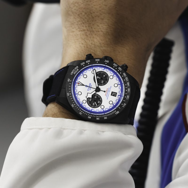 A close-up of a person's wrist wearing the M79360B-0002 watch, featuring a white face, black bezel, and black strap. The person is dressed in a white sleeve.