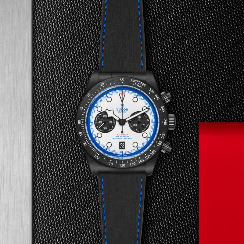 The Tudor chronograph wristwatch M79360B-0002, featuring black and white with blue accents, is displayed on a textured black surface beside a red rectangular object.