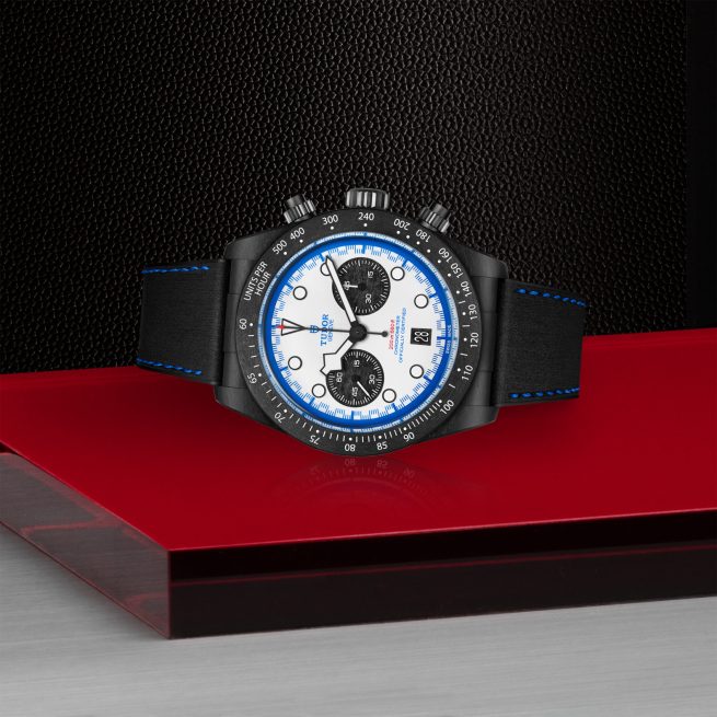 The M79360B-0002 is a black chronograph wristwatch with a white and blue face and black strap, shown on a glossy red and metallic gray surface against a textured black background.
