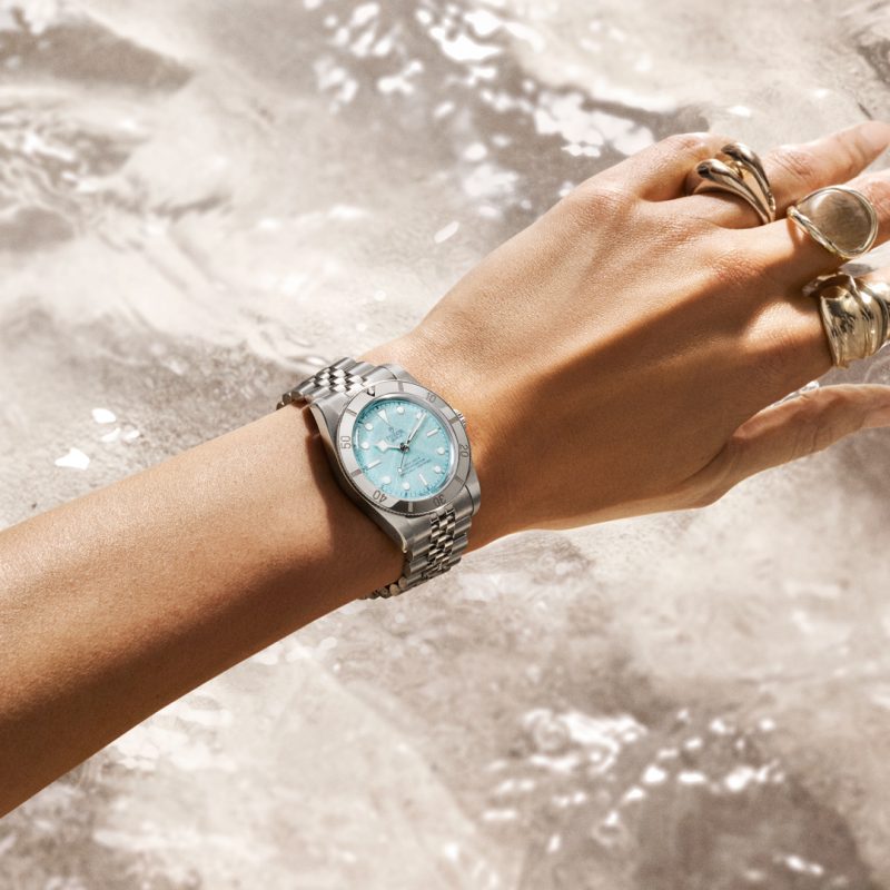 A hand adorned with multiple rings and the M79000-0001 silver wristwatch with a light blue face is displayed against rippling water.