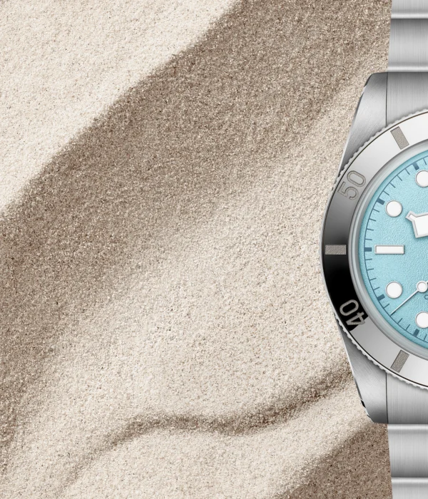 A silver TUDOR wristwatch with a light blue dial is displayed on a background of textured sand.