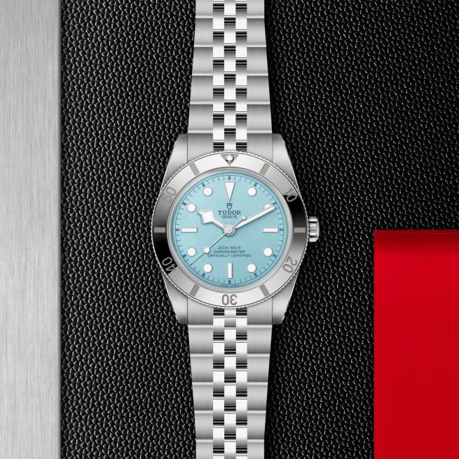 The Tudor M79000-0001, a silver wristwatch with a blue dial and metal link bracelet, is displayed on a textured black surface beside a red and silver background.