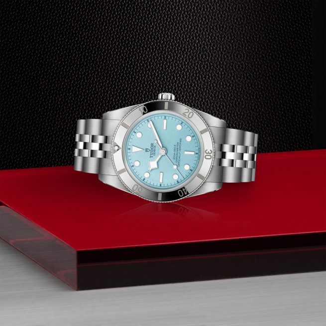 The M79000-0001, a silver wristwatch with a light blue face and metal bracelet, is displayed on a red and black geometric surface against a textured black background.