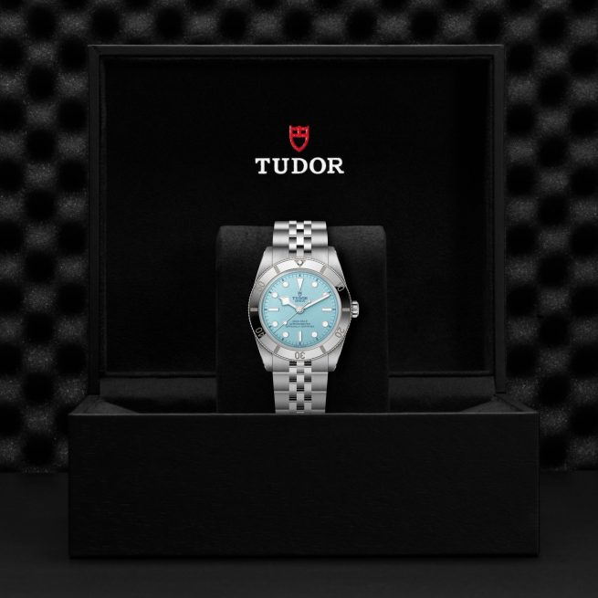 The Tudor M79000-0001 wristwatch, featuring a silver strap and light blue dial, is displayed in an open black box bearing the Tudor logo on the inside lid.