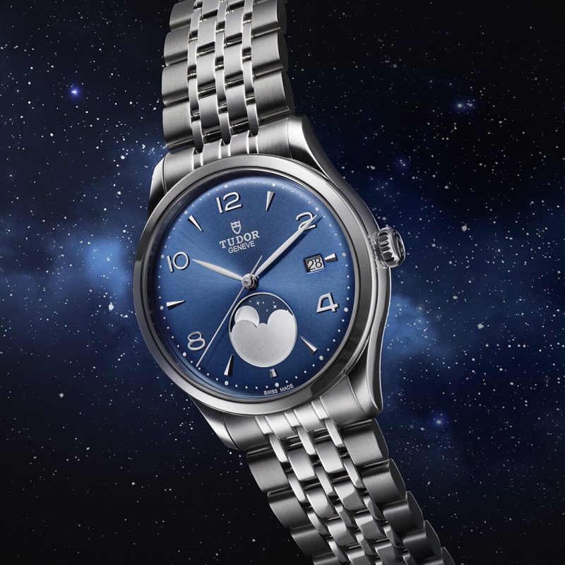 The M91560-0002 Tudor wristwatch features a blue dial, silver hour markers, and a moonphase display, set against a starry space background.