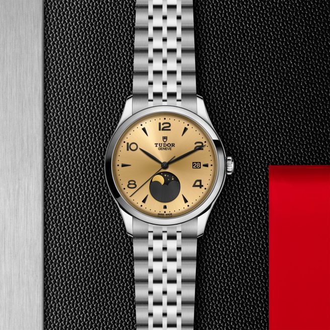 The Tudor M91560-0001 wristwatch has a silver metal bracelet and gold dial with date window and moonphase display, showcased on a textured black and silver background with a touch of red.