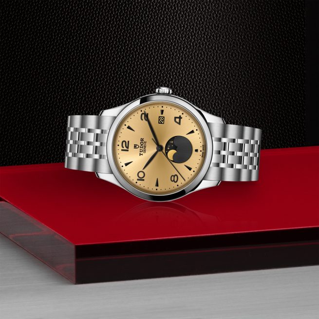 The Tudor M91560-0001 wristwatch, featuring a gold face with black numerals and a moonphase subdial, is displayed on a red and silver geometric surface against a dark textured background.