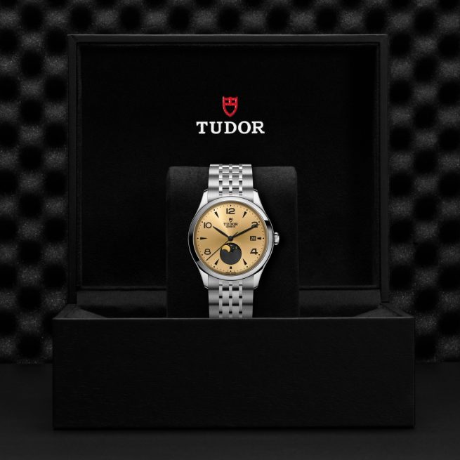 The Tudor M91560-0001 wristwatch, featuring a gold face and silver metal band, is shown in an open black Tudor box with the brand logo on the inside of the lid.