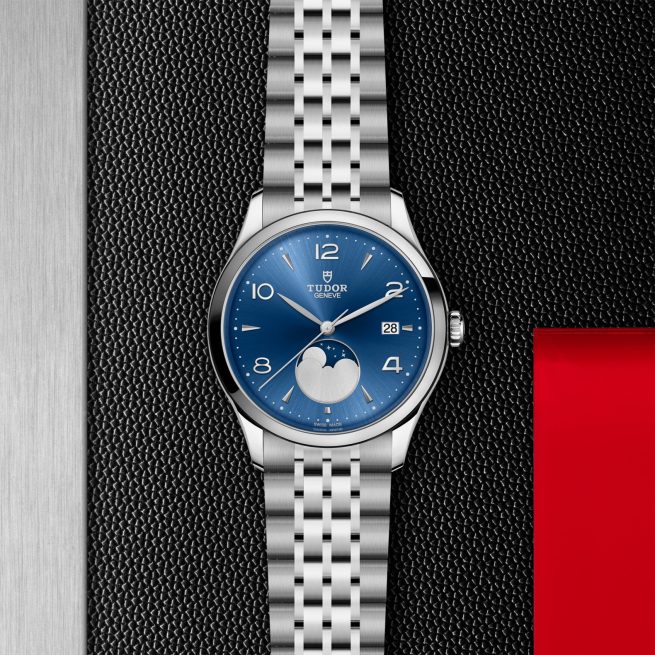 The Tudor M91560-0002 wristwatch, featuring a silver case, blue dial, moonphase display, and date window, is shown on a textured black surface beside a red and metallic background.