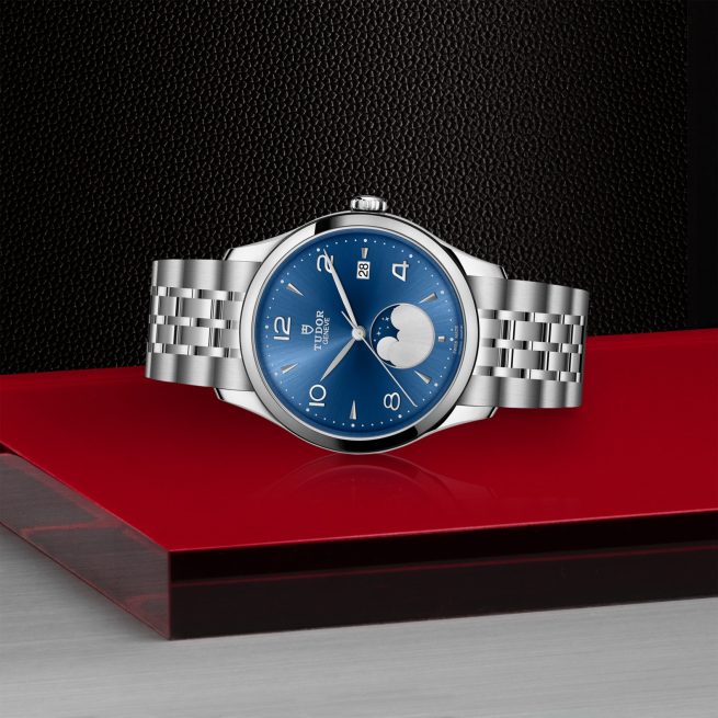 The M91560-0002, a stainless steel watch featuring a blue dial and moonphase display, is showcased on a red and silver platform against a black textured background.