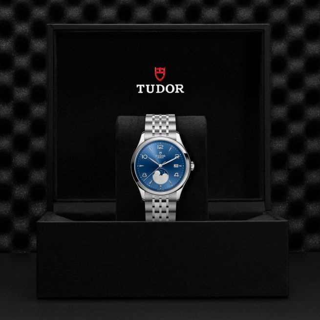 The Tudor M91560-0002 silver wristwatch with a blue dial is displayed in an open black Tudor box against a textured black background.
