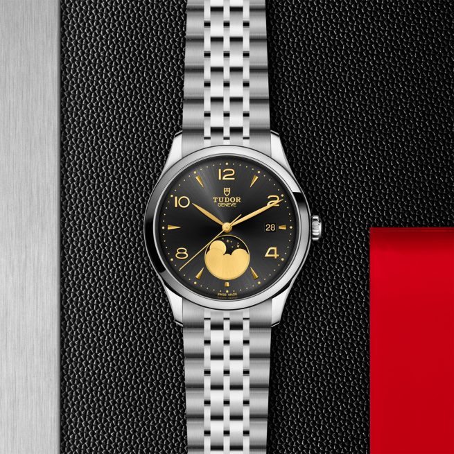 The Tudor M91560-0003 wristwatch features a silver metal band, black dial, gold moon phase display, and date window, all set against a textured black and red background.