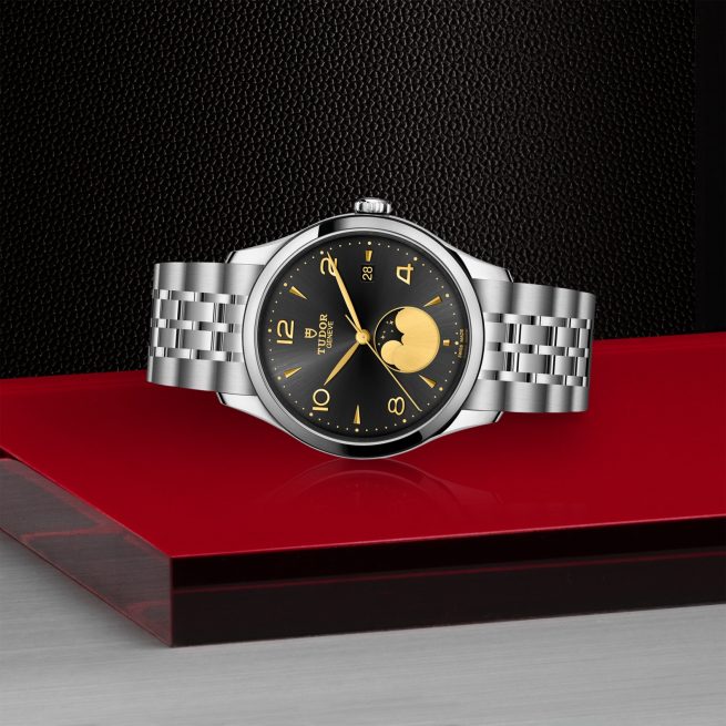 The M91560-0003 is a silver Tudor wristwatch with a black and gold dial, moon phase feature, and metal bracelet, displayed on a red and black platform.