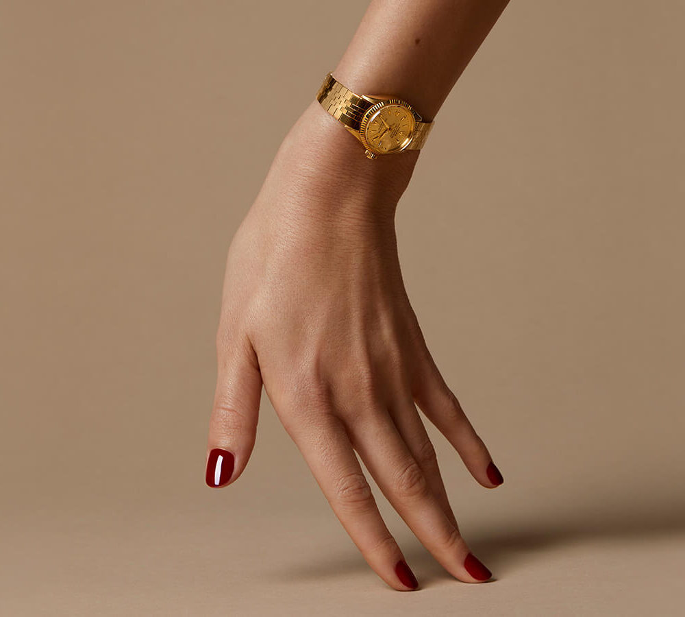 A woman's hand wearing a gold watch against a beige background, with manicured nails painted red.