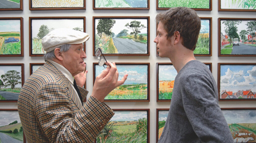 An older man in a tweed cap uses a magnifying glass to examine a younger man's face, both standing in front of a wall of landscape paintings.