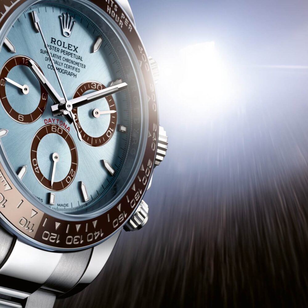 Close-up of a Rolex Daytona watch with a blue and white dial, displayed against a blurred background with a light flare.