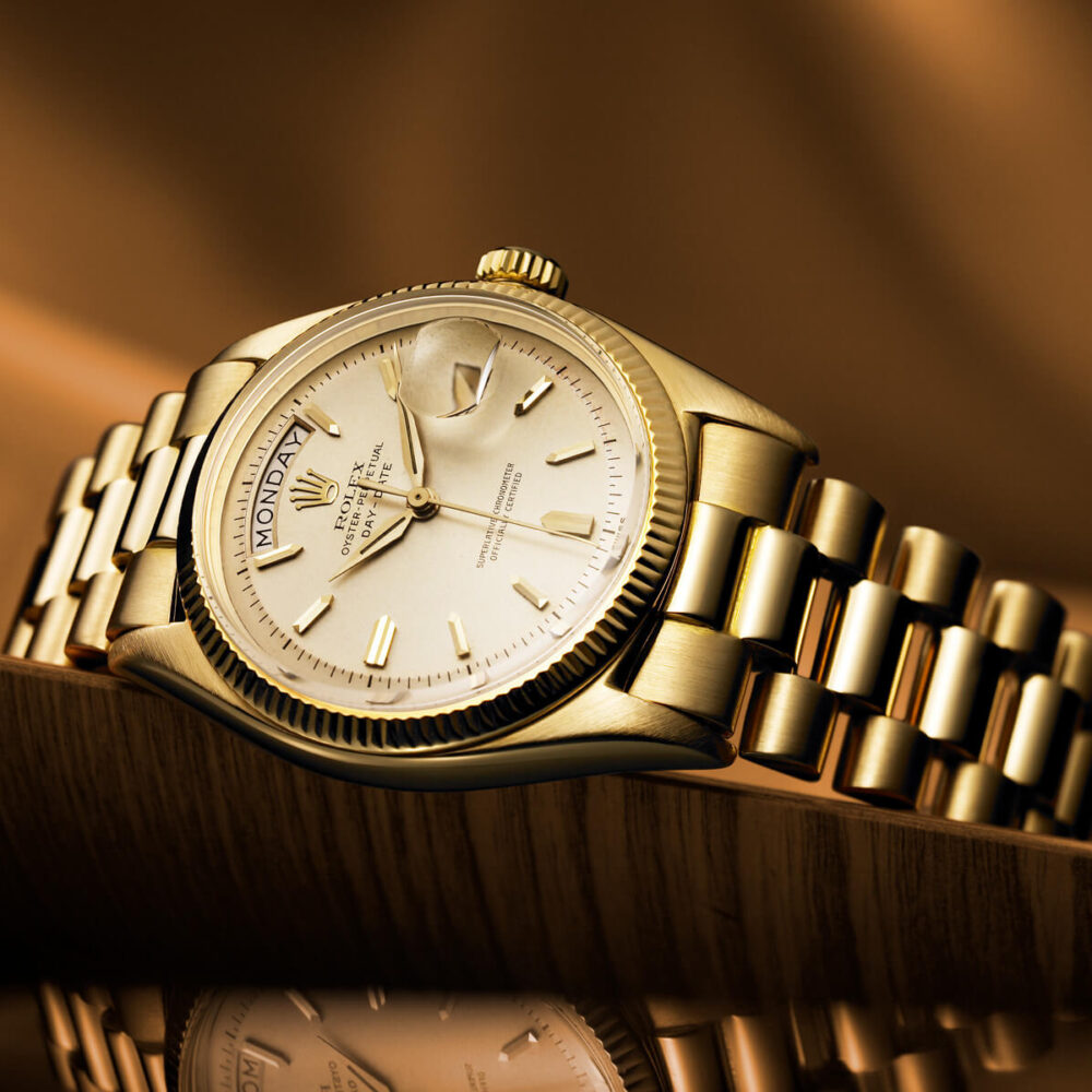 A gold Rolex watch with a date display, resting on a polished surface with its reflection visible.
