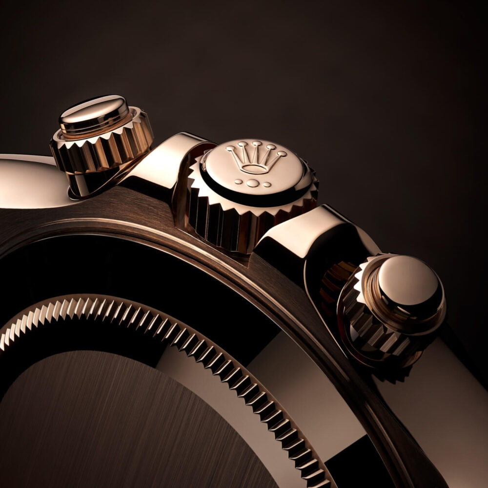 Close-up of a luxury watch showing its crown and partial dial, in polished gold and silver tones on a dark background.